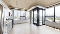 This contemporary urban residence showcases a striking industrial-modern aesthetic throughout. The main living area immediately impresses with its exposed concrete ceiling, polished visible ductwork, and structural concrete columns that lend an authentic loft-style character. Floor-to-ceiling windows wrap around the living space, flooding the interior with abundant natural light and framing expansive city skyline views. A distinctive glass-enclosed balcony vestibule with dark-framed sliding doors provides direct outdoor access while serving as a refined architectural focal point.

The kitchen is outfitted with sleek two-tone cabinetry — high-gloss white uppers paired with textured grey lowers — complemented by a white subway tile backsplash, quartz countertops, and a full suite of stainless steel appliances including a gas range, built-in microwave, and dishwasher.

The bedroom features light wood-style flooring, a dedicated walk-in closet, and direct access to a private ensuite bathroom, while exposed concrete above continues the industrial design language. Two well-appointed bathrooms serve the residence: one with a glass-enclosed walk-in shower with white subway tile and a modern vanity, and a second full bath with a tub-shower combination finished in matching tile. Light wood-style flooring flows consistently throughout, tying the clean, minimalist palette together.