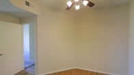 This single-floor residential space presents a clean, straightforward layout designed for comfortable everyday living. The main living area is finished with warm hardwood flooring that adds a natural, inviting tone throughout the space. Neutral beige walls provide a versatile backdrop, making it easy to personalize with furnishings and décor of any style. A ceiling fan with an integrated light fixture serves as the central overhead feature, offering both ambient lighting and practical air circulation. Crisp white baseboards frame the room neatly, complementing the overall clean aesthetic. A doorway visible from the main area leads to an adjacent room or hallway, suggesting a connected and functional floor plan. The space benefits from a simple, uncluttered design that emphasizes practicality and ease of use. The consistent flooring and wall tones carry a cohesive feel throughout, contributing to a calm and approachable atmosphere. Overall, this single-floor space offers a solid residential foundation with classic finishes and a layout that lends itself well to a variety of living arrangements.