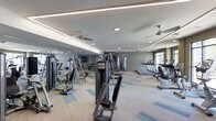 This fitness center is designed to accommodate a comprehensive workout experience within a single, open-plan floor. The space is furnished with an extensive selection of strength-training machines, including cable systems and weight stations, as well as a dedicated row of cardio equipment such as treadmills and ellipticals positioned along one side. The layout provides ample room to move between stations comfortably.

The ceiling features a combination of recessed lighting and continuous linear LED strips that run along the perimeter, creating even, well-distributed illumination throughout the space. Multiple ceiling fans are mounted overhead, supporting air circulation for an added level of comfort during workouts. A full wall of floor-to-ceiling mirrors along one side visually expands the room while allowing users to monitor their form.

The flooring consists of multi-toned carpet tiles in neutral tones accented with blue and gold, adding a contemporary aesthetic to the space. Natural light is introduced through a window on the far wall, complementing the interior lighting. The overall design is clean, modern, and functional, creating a focused environment suited for a variety of fitness routines.