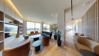 This elegant, single-floor space is defined by its open-plan layout, seamlessly connecting multiple functional zones through thoughtful architectural design. Light oak flooring flows throughout, unifying the space and complementing the warm, neutral palette accented by soft cove lighting along the ceilings.

The lounge area is anchored by a mix of velvet and leather seating arrangements — including curved sofas and accent chairs — centered around sculptural coffee tables. Vertical wooden slat partitions serve as architectural dividers, adding texture and visual interest while maintaining an open, airy feel. Lush indoor plants are positioned throughout, adding organic warmth to the contemporary setting.

The kitchen is a standout feature, showcasing expansive Calacatta marble countertops, a large central island with bar seating, and dramatic black dome pendant lights with gold interiors. Integrated cabinetry in warm wood tones and full-height marble cladding on structural columns elevate the space further.

The dining area offers an intimate setting framed by oversized black-framed windows, through which a striking large-scale artwork is displayed as a window treatment, providing both privacy and artistic impact.

Floor-to-ceiling windows throughout the space flood every zone with natural light and frame panoramic views of the surrounding cityscape and a green rooftop garden, creating a bright and inspiring atmosphere at every turn.