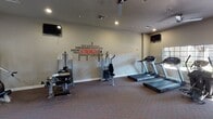 This fitness center offers a comprehensive workout environment designed to support a wide range of exercise routines. The space is laid out across a single floor and features durable patterned carpet throughout, recessed lighting, and ceiling fans that help maintain a comfortable atmosphere during workouts.

The cardio area is equipped with multiple treadmills, elliptical machines, and a stair climber, all positioned near windows that bring in natural light and provide outdoor views. A mounted flat-screen television is available for entertainment during cardio sessions.

The strength training section houses a multi-functional cable machine, selectorized weight equipment, and additional resistance machines, offering a solid variety of options for full-body strength workouts. Exercise balls are also available for supplemental training. A motivational wall decal featuring fitness-themed words such as "Strength," "Endurance," and "Reps" adds an energizing visual element to the space.

Large glass sliding doors lead to an adjacent area, expanding the usable space and allowing for flexible workout arrangements. A glass-panel exterior door provides direct outdoor access. Safety features include a mounted fire extinguisher and smoke detectors. Overall, the fitness center presents a practical, well-organized environment suited for daily exercise.