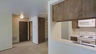This single-floor unit presents a functional and straightforward layout designed to maximize usability. The main living area features neutral carpet flooring and light-toned walls, creating a clean and understated atmosphere. A standout feature is the pass-through kitchen, which connects seamlessly to the living space and is equipped with dark wood cabinetry, a white electric range, a built-in microwave, and a sink — providing an efficient cooking and prep environment within a compact footprint.

Storage is a notable strength of this unit. An open built-in shelving unit with a clothes rod offers ample wardrobe and organizational space directly within the living area, while dark wood-paneled doors lead to additional rooms, including what appears to be a bathroom. A floor-level air vent is integrated into the living area, indicating a central heating system.

The overall aesthetic blends warm wood tones with white and neutral surfaces, giving the space a modest yet practical character. Ceiling-mounted lighting provides consistent illumination throughout. The layout flows logically, with clear sightlines from the kitchen through to the living and storage areas, making the space feel cohesive and easy to navigate.