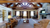 This distinguished single-story space makes an immediate impression with its dramatic vaulted ceiling, where rich, dark wood trusses and beams create a striking architectural focal point overhead. The open, airy volume of the main living area is enhanced by a row of large windows dressed in floor-length brown drapery, allowing generous natural light to fill the room while maintaining an air of warmth and sophistication.

The main gathering area is thoughtfully appointed with traditional furnishings, including plush upholstered sofas and armchairs arranged around a classic carved wood coffee table, all set atop a soft wall-to-wall carpet. The warm beige and brown tones of the walls and décor complement the deep wood accents throughout, creating a cohesive and inviting atmosphere.

A dining area is visible toward the back of the space, furnished with ornate chairs and a formal table, offering a seamless transition from the relaxed seating area. Lush potted plants are placed throughout, adding touches of natural greenery that soften the interior. The combination of exposed structural woodwork, high ceilings, abundant windows, and carefully chosen traditional furnishings gives this space a grand yet comfortable character, well-suited for both relaxed gatherings and more formal occasions.