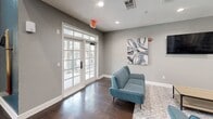 Located at 5720 Forest Park Rd in Dallas, TX, this single-floor communal space offers an impressive and welcoming environment defined by its dramatic architectural features and thoughtful layout. The main living and dining area commands attention with soaring double-height ceilings and expansive floor-to-ceiling French doors flanked by transom windows, flooding the space with abundant natural light and providing views to the outdoors. Rich, dark hardwood flooring runs throughout this open area, complementing the soft gray walls and white trim for a clean, modern aesthetic. High-top dining tables with industrial-style seating create a casual gathering atmosphere, while a dedicated lounge area features teal upholstered seating, a wall-mounted flat-screen television, and coordinating abstract wall art. A pendant chandelier and recessed lighting add warmth and versatility to the space. The fully equipped kitchen features warm honey-toned cabinetry, stone-look countertops, slate tile flooring, and stainless steel appliances including a refrigerator, range, and microwave. Track lighting overhead ensures excellent task visibility. A coffee and beverage station adds a hospitable, amenity-rich touch. A rounded tower entrance with arched windows further distinguishes the architectural character of this inviting, multi-functional space.