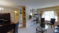 This single-floor residence offers a thoughtfully designed open-concept layout that maximizes both functionality and comfort. The living area is carpeted and bathed in natural light from a window dressed with elegant draping curtains, creating a warm and inviting atmosphere. A glass-top coffee table and seating anchor the space, lending it a classic yet practical feel.

A defining architectural feature is the built-in floor-to-ceiling bookshelf unit that serves as a partial room divider, seamlessly separating the living area from the hallway and kitchen corridor. This divider adds character while providing ample storage and display space. Framed artwork adorns the walls throughout, contributing to a curated, homely aesthetic.

The breakfast bar extends from the divider wall, offering a casual dining or work surface complemented by barstool seating. A television is mounted nearby, making this a natural media and gathering hub. The hallway visible beyond the divider leads toward the kitchen, where a full-sized refrigerator can be seen, suggesting a well-equipped culinary space. The neutral color palette of warm beige and white tones ties the entire floor together, creating a cohesive and comfortable living environment.