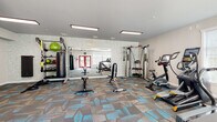 This single-floor fitness center is thoughtfully designed to support a comprehensive workout experience. The space features a modern aesthetic, highlighted by eye-catching geometric-patterned carpet in shades of gray, brown, and blue that adds visual interest throughout the room. Flush-mounted rectangular light fixtures provide consistent, bright illumination, while multiple windows dressed with white blinds invite ample natural light and views of the surrounding greenery.

The main workout area is generously sized and well-organized, housing a full array of equipment to accommodate varied fitness routines. Cardio options include multiple treadmills and elliptical machines, while a stationary cycle provides additional variety. Strength training is well-represented with a multi-station cable machine, adjustable weight benches, a dumbbell rack, and a dedicated functional training station featuring a punching bag, medicine balls, kettlebells, and resistance training accessories.

A textured accent wall with a mosaic tile pattern adds architectural character to the space. A secondary area near the entrance offers a practical amenity zone with bright blue lockers, a seating bench, a water fountain, and access to a restroom. Red double doors serve as the main entry point, contributing a bold design contrast. Overall, the fitness center combines functionality and style to create an inviting and motivating environment.