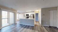 This well-appointed single-floor residence offers a thoughtfully designed open-concept layout that seamlessly connects the kitchen, dining, and living areas. The kitchen serves as the centerpiece of the space, featuring a large central island with a white countertop and built-in sink, crisp white cabinetry, and a full suite of stainless steel appliances including a refrigerator, range, and microwave. Recessed and overhead lighting keeps the kitchen bright and functional. The living area extends naturally from the kitchen and is bathed in natural light, with sliding glass doors opening to a private balcony. Warm hardwood-style flooring flows consistently throughout the main living spaces, lending a cohesive and contemporary feel. A dedicated laundry area with washer and dryer hookups is conveniently tucked adjacent to the kitchen. The bedroom offers a clean, neutral palette with the same durable hardwood-style flooring and ample wall space. The bathroom is a standout feature, boasting a full soaking tub surrounded by striking large-format marble-patterned tile in white and grey tones, complemented by brushed nickel fixtures. A separate vanity area includes a wide white countertop, under-mount sink, and a large mirror with overhead lighting. The overall ambiance is clean, modern, and move-in ready.