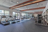 Located at 1099 Boulevard SE in Atlanta's Grant Park neighborhood, this fitness center offers a well-appointed, single-floor exercise facility designed to support a comprehensive workout experience. The space is defined by its eye-catching ceiling beams accented in bold red, orange, and teal, adding a dynamic visual energy to the otherwise clean, white interior. Large windows along the perimeter flood the room with abundant natural light and provide open views of the surrounding streetscape, creating an inviting and motivating atmosphere. The flooring throughout consists of durable, dark patterned carpet tile suited for high-traffic fitness use, complemented by rich wood base trim that adds a polished finish. The cardio zone features a row of Matrix treadmills and elliptical machines positioned along the windowed walls, maximizing the connection to natural light and outdoor views. A dedicated strength training area is equipped with a functional cable machine, a spin bike, and a selection of resistance machines and free weights, all arranged to allow ample room for movement. Full-length mirrors line one wall, enhancing the sense of spaciousness throughout. Recessed lighting and an acoustic tile ceiling complete the practical yet thoughtfully designed interior, making this fitness center a well-rounded amenity.