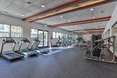 Located at 1099 Boulevard SE in Atlanta's Grant Park neighborhood, this fitness center offers a well-appointed, single-floor exercise facility designed to support a comprehensive workout experience. The space is defined by its eye-catching ceiling beams accented in bold red, orange, and teal, adding a dynamic visual energy to the otherwise clean, white interior. Large windows along the perimeter flood the room with abundant natural light and provide open views of the surrounding streetscape, creating an inviting and motivating atmosphere. The flooring throughout consists of durable, dark patterned carpet tile suited for high-traffic fitness use, complemented by rich wood base trim that adds a polished finish. The cardio zone features a row of Matrix treadmills and elliptical machines positioned along the windowed walls, maximizing the connection to natural light and outdoor views. A dedicated strength training area is equipped with a functional cable machine, a spin bike, and a selection of resistance machines and free weights, all arranged to allow ample room for movement. Full-length mirrors line one wall, enhancing the sense of spaciousness throughout. Recessed lighting and an acoustic tile ceiling complete the practical yet thoughtfully designed interior, making this fitness center a well-rounded amenity.