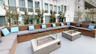 This thoughtfully designed outdoor terrace offers a versatile and inviting communal space across a single level. The terrace is organized into distinct zones, each serving a specific purpose while maintaining a cohesive aesthetic rooted in warm wood tones, concrete finishes, and lush greenery.

A covered pergola section, framed by dark steel columns and rich wood overhead beams, shelters a dedicated outdoor kitchen area equipped with a stainless steel built-in grill and prep counter finished with decorative tile. Adjacent dining tables and chairs provide ample space for al fresco meals.

Moving further into the terrace, a spacious open-air deck features a central fire pit surrounded by a curved built-in bench with cushioned seating, ideal for gathering. A mature olive tree anchors this area, providing natural shade and an organic focal point. Raised wood-clad planters throughout the space are filled with a variety of shrubs and trees, creating a sense of privacy and a connection to nature amid the urban surroundings.

The third zone extends along a wide concrete walkway flanked by tiered wood-paneled planter beds and modular lounge seating with a second fire feature. The overall ambiance is modern yet warm, blending natural materials with clean architectural lines for a polished and functional outdoor retreat.