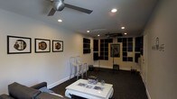 This single-floor space is thoughtfully designed as a combined music studio and lounge, offering a functional yet comfortable environment for creative work. The room features extensive acoustic treatment panels mounted across multiple walls, arranged in a grid pattern to optimize sound absorption and reduce unwanted reflections — a clear nod to its purpose as a serious audio environment. Recessed lighting is distributed evenly across the smooth white ceiling, providing consistent, adjustable illumination throughout, while ceiling fans ensure comfortable airflow during long sessions. The studio side of the space is equipped with microphone stands and amplifiers, ready for recording or live practice. Framed music-themed artwork adorns the walls, adding a personal and artistic character to the room. A lounge area anchors the opposite side, featuring a white coffee table and a plush sofa, creating a relaxed space for listening, collaborating, or unwinding between sessions. Dark carpeting runs throughout, further contributing to the room's acoustic properties while lending a sleek, professional aesthetic. Together, the design elements create a well-balanced space that merges the practicality of a recording environment with the comfort of a creative retreat.