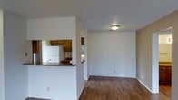This single-floor residential space offers a practical and functional layout designed for comfortable everyday living. The open-concept design connects the living area to the kitchen through a half-wall pass-through, creating a sense of visual continuity while maintaining defined zones for cooking and relaxing. The living area features warm hardwood-style flooring that runs throughout, adding a cohesive and inviting feel to the space. A flush-mount ceiling light provides central illumination, complementing the textured ceiling that extends across the unit. The kitchen is equipped with classic wooden cabinetry in a warm brown finish, a white refrigerator, and a dark laminate countertop that wraps along the pass-through bar, offering additional prep or dining space. A secondary hallway visible from the main living area leads to additional rooms, suggesting a well-organized floor plan. The clean white walls throughout the space provide a neutral backdrop, making it easy to personalize with furnishings and décor. Overall, the space presents a straightforward, move-in-ready layout that balances openness with functional room separation.