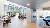 Located at 7111 East Tierra Buena Lane in Scottsdale, Arizona, this sophisticated community living space delivers a striking first impression through its dramatic double-height ceilings and expansive floor-to-ceiling glass curtain walls. Flooded with abundant natural light, the single-floor layout offers generous, open-plan proportions finished with light-toned hardwood flooring that runs seamlessly throughout.

A sculptural marble-clad fireplace serves as a bold architectural centerpiece, dividing the space while anchoring distinct seating areas on either side. The main lounge features oversized sectional sofas, plush accent chairs, and curated greenery positioned to take full advantage of panoramic views of the surrounding Scottsdale streetscape. Elegant pendant chandeliers and contemporary linear fixtures provide sophisticated ambient lighting above.

A dedicated dining area offers built-in bench seating alongside café-style tables and chairs, while a long communal bar-height table with navy blue seating provides additional gathering space. A recreation zone with a billiards table adds a leisure-focused dimension to the layout. Steel-framed glass doors provide a visual connection between zones and lead to exterior access points. Large-scale artwork and carefully chosen decorative accessories complete the refined, modern aesthetic of this well-appointed community gathering space.