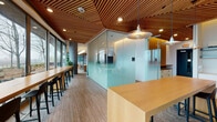 Located near the iconic Burnside Bridge in Portland, Oregon, this thoughtfully designed single-floor workspace combines natural materials, modern architecture, and abundant natural light to create a refined and functional environment. A signature sculptural wood slat ceiling runs throughout the space, adding warmth and acoustic character while lending a distinctly organic quality to the interior.

Floor-to-ceiling windows line the perimeter, framing panoramic views of the Willamette River, surrounding greenery, and the Portland skyline. These expansive glazed walls flood the space with natural light throughout the day. Exposed concrete columns punctuate the open layout, complementing the raw, industrial undertones that contrast beautifully with the warm wood tones and soft textile furnishings.

The workspace is organized into several distinct zones. A central open area features long communal tables with bar-height seating beneath pendant lights, ideal for collaborative work. Glass-partitioned meeting rooms with frosted panels offer privacy while maintaining visual connectivity. A smaller conference room with a round table and wall-mounted display serves focused gatherings, while a larger meeting room with floor-to-ceiling windows overlooks the river.

A compact, minimalist kitchenette with clean white cabinetry and cork flooring supports day-to-day needs. Lounge areas with plush navy sofas, accent chairs, and lush indoor plants are positioned alongside vertical wood slat walls, creating inviting spaces for informal interaction or quiet reflection.