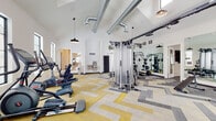 This single-story amenity building is designed with a clean, modern aesthetic and thoughtful layout. The entrance lobby greets visitors with soaring vaulted ceilings, light wood-look flooring, and a striking circular chandelier with exposed bulbs that adds a warm, industrial-inspired touch. Large black-framed glass doors invite natural light into the space, while wide corridors connect seamlessly to various amenity areas.

The mailroom is a standout feature, showcasing the same soaring ceiling height and an elaborate chandelier overhead. Multiple banks of individual mailboxes line the walls and central island, which also incorporates built-in cabinetry with wood-toned finishes and a white countertop. A corkboard bulletin board keeps residents informed.

The fitness center is expansive and impressively equipped, featuring vaulted ceilings with exposed silver ductwork and rows of clerestory windows that flood the space with natural light. Pendant lights hang from above, complementing the bold geometric yellow, grey, and white patterned carpet below. The gym houses a full array of equipment including cable machines, free weights, benches, Echelon spin bikes, ellipticals, and a heavy bag. Full-length mirrors along one wall expand the sense of space and enhance the workout environment.