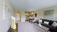This single-floor unit showcases an open-concept design that seamlessly connects the living, dining, and kitchen areas into one cohesive space. The living area features plush, light-toned carpeting and is furnished with a dark upholstered sofa, a gray coffee table, and decorative yellow accents that add warmth throughout. Adjacent to the living space, a compact dining area accommodates a dark wood table with white upholstered chairs, ideal for everyday meals or casual gatherings.

The kitchen is situated at the rear of the open layout and features white cabinetry, white appliances, and a peninsula counter with bar seating that acts as a natural divider between the kitchen and dining areas. A pendant light hangs above the peninsula, while a ceiling fan provides additional airflow. The kitchen has tile flooring that transitions into the carpeted living and dining zones.

The entryway, visible from the main living space, offers a clean and straightforward passage into the unit. Neutral beige walls and a textured ceiling run throughout, contributing to the space's cohesive, understated aesthetic. Overall, the unit presents a functional and well-organized layout with a warm, inviting ambiance.