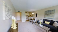 This single-floor unit showcases an open-concept design that seamlessly connects the living, dining, and kitchen areas into one cohesive space. The living area features plush, light-toned carpeting and is furnished with a dark upholstered sofa, a gray coffee table, and decorative yellow accents that add warmth throughout. Adjacent to the living space, a compact dining area accommodates a dark wood table with white upholstered chairs, ideal for everyday meals or casual gatherings.

The kitchen is situated at the rear of the open layout and features white cabinetry, white appliances, and a peninsula counter with bar seating that acts as a natural divider between the kitchen and dining areas. A pendant light hangs above the peninsula, while a ceiling fan provides additional airflow. The kitchen has tile flooring that transitions into the carpeted living and dining zones.

The entryway, visible from the main living space, offers a clean and straightforward passage into the unit. Neutral beige walls and a textured ceiling run throughout, contributing to the space's cohesive, understated aesthetic. Overall, the unit presents a functional and well-organized layout with a warm, inviting ambiance.