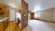 This single-floor unit offers a practical and straightforward layout with a warm, classic character defined by its distinctive wood-paneled accent walls. The kitchen is efficiently designed as a galley-style space, featuring warm wood cabinetry, light stone-finish countertops with a stainless sink, and white appliances including a range and refrigerator, all set on easy-care laminate hardwood flooring. A striking floor-to-ceiling wood-paneled partition wall separates the kitchen area from the main living space, adding a natural, rustic element to the interior. The living area is open and generously proportioned, with carpeted flooring, neutral-toned walls, and a window that brings in natural light and views of the surrounding greenery. A wall-mounted HVAC unit provides climate control. The bedroom is a calm, carpeted retreat featuring a large sliding window that frames outdoor views and fills the room with ample natural light. The bathroom is clean and functional, equipped with a tub and shower combination, a white vanity with storage, and a mirrored medicine cabinet above, all finished with warm laminate wood flooring. Wood-tone doors and trim details carry a consistent, cohesive aesthetic throughout the entire space.