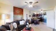 This single-floor residence showcases an open-concept layout that connects the living area and kitchen in one cohesive, flowing space. The living room is anchored by a plush, dark sectional sofa adorned with decorative pillows, positioned beneath a classic ceiling fan with integrated lighting. A striking large-scale abstract wall piece in warm amber and earth tones serves as a bold focal point, complementing the room's warm, eclectic aesthetic. Decorative accents, including a sunburst mirror and sculptural giraffe figurine, add personality and visual interest throughout.

The kitchen flows naturally from the living area and features dark, rich cabinetry paired with a granite-topped breakfast bar, providing both workspace and casual seating via bar stools. Black appliances, including a range and refrigerator, provide a cohesive, contemporary look. The kitchen is well-lit with under-cabinet and overhead lighting that enhances the richness of the cabinetry. Light-toned carpeting runs throughout the living space, softening the overall palette and adding comfort underfoot. The space is well-lit with ceiling fans in both zones, creating a comfortable and stylish environment.