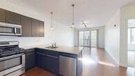 Located at 163 Franklin Street in downtown Stamford, CT, this well-appointed one-bedroom, one-bathroom residence offers a thoughtfully designed open-concept layout across a single floor. The kitchen serves as a central hub of the space, featuring dark espresso cabinetry, granite countertops, a peninsula island with a built-in sink, stainless steel appliances including a refrigerator, gas range, microwave, and dishwasher, and stylish pendant lighting overhead. The kitchen flows seamlessly into the living area, which is bathed in natural light from large sliding glass doors that open to a balcony with neighborhood views. Warm hardwood flooring extends throughout the main living and kitchen areas, adding a polished, cohesive feel. The generously sized bedroom is finished with plush neutral carpeting and features a window that draws in additional natural light. A dedicated closet provides convenient storage. The bathroom is cleanly designed with tile flooring, a white vanity with ample cabinet space, a large mirror, and crisp white finishes throughout. A hallway connects the private areas of the home, completing the functional and modern floor plan. The overall ambiance is bright, clean, and contemporary.