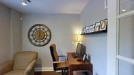 This single-floor space offers a welcoming and well-appointed home office environment designed for both productivity and comfort. The workspace is anchored by a rich wooden desk paired with an ergonomic high-back chair, providing a practical and comfortable area for work or study. A desktop computer setup, keyboard, and printer are neatly arranged, reflecting a fully functional office configuration. The walls are finished in a soft, neutral gray tone that contributes to a calm and focused atmosphere. A large, ornate round wall clock with an intricate wrought-iron frame serves as a striking decorative focal point, adding a touch of classic elegance to the space. A floating display shelf runs along one wall, showcasing a series of framed letter prints that add a personalized and creative flair. Warm ambient lighting from a decorative table lamp with a floral motif casts a soft glow, enhancing the inviting character of the room. A glimpse of a cozy upholstered seating piece in the corner suggests the space seamlessly blends work functionality with relaxed comfort. The clean lines, neutral palette, and thoughtful decor choices come together to create a space that is both organized and aesthetically pleasing.