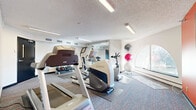 This fitness center offers a practical and well-organized space designed to support a range of exercise routines. The room is laid out efficiently, making good use of its footprint to accommodate a variety of equipment without feeling cramped. Flooring consists of patterned commercial carpet with protective rubber mats positioned beneath each piece of equipment.

The cardio section is well-stocked, featuring Matrix-brand treadmills, elliptical trainers, and upright stationary bikes, all arranged to allow comfortable use. A dumbbell rack with a selection of free weights lines one wall, providing options for strength training. Stability balls in varying sizes are neatly stored on a vertical rack near a large arched window, which allows natural light to fill the space and offers a pleasant view outward.

A full-length wall mirror spans a significant portion of one wall, visually expanding the room and allowing users to monitor their form during workouts. A bold red accent wall adds a dynamic, energizing element to the otherwise neutral white and grey palette. A wall-mounted flat-screen television is positioned on the red wall for entertainment during workouts.

Overhead fluorescent strip lighting provides consistent, even illumination throughout the space. A textured ceiling and standard HVAC venting complete the functional interior. The room is accessed through a solid door with a decorative window insert.