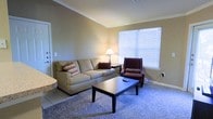 This single-floor space offers a welcoming and functional layout designed for comfortable everyday living. The living area serves as the centerpiece of the space, featuring warm neutral-toned walls accented by classic crown molding that adds a refined architectural touch. A large window dressed with white blinds allows generous natural light to fill the room, creating a bright and inviting atmosphere.

The living area is finished with plush carpeting layered over tile flooring near the entryway, providing both comfort and practicality. The space is furnished with a spacious beige sofa accented by decorative throw pillows, complemented by a deep burgundy accent chair, and anchored by a dark wood rectangular coffee table — together creating a cohesive and cozy seating arrangement. A classic table lamp adds warm ambient lighting, enhancing the relaxed feel of the room.

The layout flows naturally, with the transition between the tiled entryway and carpeted living area subtly defining distinct functional zones. The overall ambiance of the space is calm, neutral, and well-lit, making it a practical and comfortable environment suitable for relaxing or entertaining.