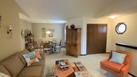 This single-floor residence offers an open and functional layout that seamlessly connects the living and dining areas into one cohesive space. The living area is comfortably appointed with a plush sofa, colorful accent rugs, and a central coffee table, creating a relaxed and inviting atmosphere. A warm-toned armoire serves as a natural divider between the living and dining zones, adding both storage and visual interest to the room. The dining area features a classic wrought-iron table and chair set, illuminated by a pendant light overhead, making it an ideal spot for everyday meals or entertaining. Light neutral walls and carpeted flooring run throughout the space, contributing to a calm and cohesive aesthetic. A set of large wooden closet doors along one wall provides ample built-in storage, keeping the space tidy and organized. Decorative wall art and thoughtful personal touches lend the space a lived-in, homey character. The layout is efficient and well-proportioned, with clear sightlines across the entire floor plan, making the space feel open and easy to navigate.