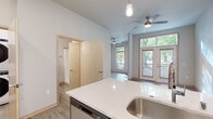 This thoughtfully designed single-floor unit offers a seamless open-concept layout that connects the kitchen and living area into one cohesive, airy space. The kitchen serves as a central hub, anchored by a large island with a crisp white quartz countertop, an undermount stainless steel sink, and elegant pendant lighting overhead. Dark espresso cabinetry provides ample storage and a rich contrast against the light finishes, while a full suite of stainless steel appliances — including a refrigerator, range, microwave, and dishwasher — completes the modern culinary space. Durable hardwood-style flooring flows throughout the entire unit, lending warmth and visual continuity. The living area benefits from soaring vaulted ceilings and a ceiling fan, while a set of French doors framed by a transom window invites natural light to fill the room and offers a pleasant view of surrounding greenery. A dedicated in-unit laundry closet houses a stacked washer and dryer, adding everyday convenience. The bathroom features a clean, contemporary design with a white countertop vanity, dark cabinetry, a large mirror, and a combination shower and tub. Neutral wall tones and clean lines throughout create a bright, welcoming atmosphere well-suited to modern living.
