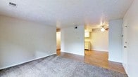 This single-floor residence offers a practical and functional open-concept layout designed to maximize usable living space. The main living area is finished with wall-to-wall carpet, providing a comfortable and warm underfoot feel, while the adjacent dining and kitchen zones transition to smooth hardwood-style flooring for easy maintenance and a clean aesthetic contrast. A textured ceiling runs throughout, adding a classic residential character to the space. The kitchen area is positioned in an open, accessible corner of the layout, featuring light-toned cabinetry and countertops that contribute to a bright, functional workspace. A ceiling fan is installed in the dining or kitchen area, promoting air circulation and adding a modest decorative touch. A partial dividing wall separates the living and kitchen spaces while maintaining the open flow of the floor plan. A hallway visible from the main living area leads to additional rooms, suggesting a well-organized layout with clearly defined zones. Neutral wall tones throughout create a versatile backdrop suitable for a variety of interior styles. The space is well-lit and benefits from a clean, move-in ready condition, making it a straightforward and comfortable single-floor living environment.