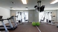 This single-level fitness room provides a practical and well-equipped space for residents or guests looking to maintain an active lifestyle. The room features a treadmill for cardiovascular exercise alongside a multi-station cable weight machine, catering to both cardio and strength training needs. Two green stability balls rest on the carpeted floor, adding versatility for core and balance exercises.

A full-length wall mirror runs along one side of the room, visually expanding the space and allowing users to monitor their form during workouts. Ceiling-mounted flat-screen televisions are positioned for easy viewing from multiple workout stations, enhancing the exercise experience.

Natural light enters through two windows fitted with blinds, while recessed and flush-mounted ceiling lights ensure consistent illumination throughout. The walls are finished in a neutral sage green tone, contributing to a calm and focused workout atmosphere. Informational notices are posted on the walls, providing guidance for users. The carpeted flooring offers comfort underfoot, making the room a functional and accessible fitness facility.