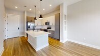 This 560-square-foot, one-bedroom, one-bathroom apartment in Charlotte, NC (Unit 160, 28206) delivers a clean, modern aesthetic throughout its single-floor layout. Light-toned hardwood flooring flows seamlessly across every room, complemented by crisp white walls, white baseboards, and matte black hardware accents that create a cohesive contemporary feel.

The kitchen is a standout feature, boasting an open-concept design with a large white waterfall island with an undermount sink, two-tone cabinetry pairing white upper cabinets with gray lower cabinets, a white subway tile backsplash, and stainless steel appliances including a refrigerator, range, and microwave. Recessed lighting and pendant fixtures illuminate the space effectively.

The 110-square-foot bedroom is bright and airy, featuring two windows with blinds that invite natural light and offer outdoor views, creating a welcoming and restful environment.

The bathroom continues the gray-and-white design language with a gray vanity, white countertop, framed mirror, and matte black fixtures. A dedicated closet accessible from the hallway provides practical storage. The overall ambiance of this unit is modern, clean, and efficiently designed for comfortable urban living.