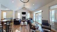 This single-floor community space is designed to foster social interaction and comfort through a series of well-appointed, open areas. The centerpiece is a spacious great room distinguished by soaring vaulted ceilings and a striking wagon-wheel chandelier that anchors the space visually. Rich hardwood-style flooring flows throughout, complementing the neutral wall tones and creating a warm, cohesive aesthetic. A stone-surround fireplace topped with a wall-mounted television serves as the focal point, flanked by large windows and glass doors that invite abundant natural light and outdoor views. Two tufted navy sofas and coordinating armchairs are arranged to encourage conversation, while white tufted ottomans add casual versatility.

Adjacent to the main lounge is a secondary seating nook with wood-framed chairs gathered around a petite round table, offering a quieter setting for small groups. Nearby, high-top pub tables with industrial-style stools provide a casual dining or gathering option. The kitchen area is equipped with warm wood cabinetry, granite countertops, black appliances including a full range and refrigerator, and a peninsula bar with bar stools — well-suited for catering or communal use. Together, these zones create a functional and inviting shared environment.