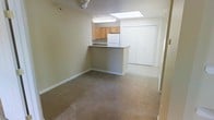 This single-floor space offers a functional and open layout that seamlessly connects the living area to the kitchen. The living space is finished with wall-to-wall carpeting in a neutral tan tone, providing a warm and comfortable feel throughout. A ceiling light fixture adds ambient illumination to the main area.

The kitchen is positioned at a slightly elevated counter height and features a peninsula-style breakfast bar that separates it from the living space while maintaining an open sightline. Natural light filters into the kitchen through a skylight overhead, brightening the area considerably. Honey-toned wood cabinetry lines the upper portion of the kitchen wall, offering ample storage, while a white refrigerator anchors the space. The countertops are finished in a neutral gray laminate.

Adjacent to the kitchen, a large set of double bi-fold closet doors provides generous storage capacity, a practical and notable feature of this unit. The walls throughout are painted in a soft, neutral beige, contributing to a clean and understated aesthetic. The overall layout is efficient and well-organized, making good use of the available floor space while keeping the environment open and uncluttered.