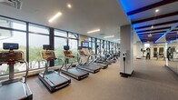 This fitness center is a comprehensively outfitted facility designed to support a wide range of workout routines. The space is organized into distinct functional zones, each tailored to a specific type of training. The cardio area runs along a floor-to-ceiling glass wall that provides abundant natural light and views of lush outdoor greenery, housing a long row of treadmills and elliptical machines, each equipped with individual screens for entertainment. Overhead, recessed linear lighting and blue accent lighting set into the ceiling create a modern, energetic atmosphere throughout.

The strength training zone features a well-stocked dumbbell rack, cable machines, benches, and a power rack with weight plates, all positioned in front of full-length mirrors that enhance the sense of space. Exposed wooden beams accented with blue lighting add an architectural contrast to the otherwise contemporary interior. A dedicated functional training area is finished with bright yellow-green flooring and is equipped with plyometric boxes, battle ropes, resistance bands, kettlebells, medicine balls, and TRX suspension trainers stored neatly on a multi-tier rack. Additional equipment, including a pull-up and dip station, occupies a quieter section of the facility. The flooring throughout is durable rubber, suitable for high-intensity use.