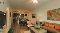 This single-floor space showcases an open-concept layout that effortlessly connects the living room, dining area, and kitchen into one cohesive, functional environment. Rich hardwood flooring flows throughout, anchoring the warm and inviting ambiance of the space.

The living area is generously sized, featuring a ceiling fan that adds both comfort and a casual charm. It is furnished with a plush sofa accented by bold orange and earth-toned throw pillows, complemented by a large ottoman that doubles as a coffee table — ideal for relaxed everyday living.

Adjacent to the living room, the dining area accommodates a full dining set, creating a natural transition between the casual lounge space and the kitchen. Pendant lighting above the dining area adds a touch of sophistication and warmth.

The kitchen is thoughtfully designed with dark cabinetry that creates a striking contrast against stainless steel appliances, including a refrigerator and microwave. A mosaic tile backsplash adds a decorative element, while track lighting ensures the workspace is well-illuminated. A kitchen island or counter bar provides additional prep space and casual seating opportunities.

Overall, the space delivers a modern yet cozy atmosphere, with a cohesive warm color palette and an efficient, open layout well-suited for both daily living and entertaining.