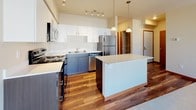 Located at 1688 Stella Avenue in Wenatchee, Washington, this single-floor residence offers a clean, modern aesthetic with a thoughtfully designed open-concept layout. The kitchen is a standout feature, showcasing two-tone cabinetry with crisp white upper cabinets and dark lower cabinets, a large central island with quartz countertops, stainless steel appliances including a refrigerator, range, and microwave, and warm hardwood flooring. Elegant pendant lighting hangs above the island, complemented by a track lighting system overhead, creating a bright and functional cooking space. The kitchen flows seamlessly into the living area, which transitions to plush carpeting and offers sliding door access to the outdoors, inviting natural light deep into the interior. The vaulted ceiling in the living space adds a sense of openness and volume. Bedrooms are carpeted and well-lit by natural light through windows that offer views of the surrounding neighborhood. The bathroom features hardwood-style flooring, a white vanity with solid countertops, and a clean, minimal design with a small window for natural ventilation. Warm wood-toned interior doors and trim throughout the home add a cohesive, inviting character to the overall space.