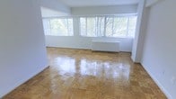 This single-floor space offers a clean, well-maintained interior defined by its standout classic parquet hardwood flooring, which adds warmth and timeless character throughout. The main living area is generously proportioned, with an open layout that allows for flexible furniture arrangement and easy movement through the space.

A row of large windows spans much of the exterior-facing wall, flooding the interior with abundant natural light and framing pleasant views of mature trees and lush greenery outside. White horizontal blinds on each window provide adjustable light control while maintaining a clean, uniform aesthetic.

The walls are finished in a crisp, neutral white, creating a bright and airy atmosphere that complements the warm tones of the hardwood floor below. A built-in baseboard heating unit is positioned along the windowed wall, ensuring comfortable climate control integrated seamlessly into the room's design.

The overall ambiance is understated and functional, with a light-filled, open character that makes the space feel welcoming and versatile. The combination of natural light, classic flooring, and neutral finishes provides a strong foundation suitable for a variety of uses and interior styles.