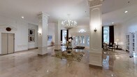 This distinguished space makes an immediate impression with its grand, light-filled lobby that sets the tone for the entire interior. The entrance hall is anchored by a series of stately white Corinthian columns with intricate carved capitals, framing the space with a sense of classical formality and architectural refinement. Underfoot, expansive polished marble flooring in warm neutral tones reflects light beautifully, enhancing the overall sense of openness and grandeur.

At the heart of the lobby, an ornate crystal and candle-style chandelier cascades from the ceiling, serving as a striking focal point. A decorative round glass table topped with an elegant floral arrangement sits atop a traditional patterned area rug, adding warmth and a welcoming touch to the space.

Natural light pours in through a series of tall glass-paneled French doors and an arched transom window at the entrance, creating a bright and airy atmosphere while offering views of lush greenery beyond. Crown molding and wainscoting detail the crisp white walls, which are accented by carefully selected artwork and wall sconces that provide soft ambient lighting.

The overall ambiance is one of timeless elegance — a thoughtfully designed space that balances grandeur with a welcoming warmth, suited for upscale gatherings or refined daily living.
