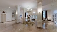 This distinguished space makes an immediate impression with its grand, light-filled lobby that sets the tone for the entire interior. The entrance hall is anchored by a series of stately white Corinthian columns with intricate carved capitals, framing the space with a sense of classical formality and architectural refinement. Underfoot, expansive polished marble flooring in warm neutral tones reflects light beautifully, enhancing the overall sense of openness and grandeur.

At the heart of the lobby, an ornate crystal and candle-style chandelier cascades from the ceiling, serving as a striking focal point. A decorative round glass table topped with an elegant floral arrangement sits atop a traditional patterned area rug, adding warmth and a welcoming touch to the space.

Natural light pours in through a series of tall glass-paneled French doors and an arched transom window at the entrance, creating a bright and airy atmosphere while offering views of lush greenery beyond. Crown molding and wainscoting detail the crisp white walls, which are accented by carefully selected artwork and wall sconces that provide soft ambient lighting.

The overall ambiance is one of timeless elegance — a thoughtfully designed space that balances grandeur with a welcoming warmth, suited for upscale gatherings or refined daily living.