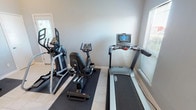 This dedicated fitness room is thoughtfully designed to support a comprehensive workout routine within a single, well-organized space. The room features light-toned tile flooring throughout, complemented by neutral gray walls and clean white trim, creating a bright and functional environment. Rubber floor mats are positioned beneath the equipment to protect the flooring and provide stability during exercise.

The cardio area is equipped with multiple machines, including a treadmill with a digital display console, an elliptical trainer, and a stationary recumbent bike, offering a range of cardiovascular training options. Natural light enters the room through windows fitted with blinds, enhancing the overall atmosphere without creating glare on equipment screens.

The strength training area features a fully stocked dumbbell rack with a wide range of weights organized in ascending order, paired with an adjustable weight bench suitable for a variety of exercises. The ceiling-mounted fan in one section of the room aids in air circulation during workouts.

Access to the room is provided through multiple entry points, including a set of French doors and a solid exterior door with a window, offering flexibility and natural ventilation. Recessed lighting and standard overhead fixtures ensure adequate illumination throughout.