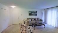 This single-floor space presents a welcoming living room designed with a neutral and understated aesthetic. The room features wall-to-wall carpeting that adds warmth underfoot, while the white walls and smooth ceiling create a bright, open atmosphere. A front entry door is positioned along one wall, providing a clear sense of entry and flow into the living area. The seating arrangement centers around a grey upholstered sofa accented with patterned throw pillows, complemented by a bold floral-patterned accent chair that adds visual interest. A dark wood coffee table with ornate legs serves as the focal point of the seating area, grounding the space with a classic touch. A tall floor lamp in the corner provides ambient lighting, supplementing the overhead light source. Wall décor includes a nature-inspired landscape canvas print, adding depth and a subtle outdoor element to the interior. The overall ambiance is calm and functional, with a straightforward layout that maximizes the usable living area while maintaining a tidy, comfortable feel suited for everyday living.