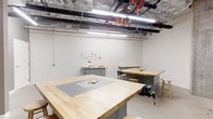This single-floor workshop studio is designed for hands-on creative and fabrication work, offering a well-organized, functional environment suited to a range of craft and making activities. The space is anchored by multiple large butcher block worktables with sage green steel bases, providing generous work surfaces for projects of varying scales. One of the central tables is equipped with a mounted bench vise, indicating suitability for more detailed fabrication or woodworking tasks. Round wooden stools are positioned throughout for comfortable seated work. The ceiling is left exposed, revealing an industrial network of HVAC ductwork wrapped in reflective insulation, black conduit, and prominent red-painted utility pipes — all of which contribute to the space's utilitarian, makerspace character. Bright fluorescent tube lighting runs along the ceiling, ensuring strong, even illumination across all work surfaces. The walls are painted white, keeping the space clean and visually open. Along the back wall, a low storage cabinet with pegboard and pinned reference materials provides practical organizational support. A cutting mat and basic tools are visible on the main table, suggesting active, ongoing use. Overall, this space delivers a practical, well-lit, and efficiently arranged environment for workshops, studio courses, or collaborative making.