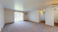 Located at 23675 Delmere Dr in North Olmsted, Ohio, this single-floor unit offers a straightforward, functional layout with several notable features throughout. The living area is generously proportioned, finished with neutral-toned carpet and bright white walls that contribute to an open, airy atmosphere. A large sliding glass door fitted with vertical blinds allows abundant natural light to fill the space while providing direct access to an outdoor view. A wall-mounted air conditioning unit and baseboard heating ensure year-round climate control. The layout, visible in the floor plan overview, reveals a well-organized arrangement connecting the living space to the kitchen and bedroom areas via a carpeted hallway. The kitchen is equipped with warm-toned wooden cabinetry, granite-style countertops, and white appliances including an electric range and full-size refrigerator, all set over wood-look vinyl flooring. The bathroom features a rich cherry wood vanity with a white countertop, a mirrored medicine cabinet, a tiled shower-tub combination, and matching wood-look flooring. The bedroom areas are carpeted and well-lit, with access to closet space and natural light provided through a window with baseboard heating beneath. Overall, the unit presents a clean, practical living environment with consistent finishes throughout.