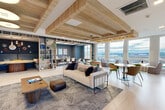 Located atop 305 Winter St in Waltham, MA, the Social Lounge Sky Deck is a thoughtfully designed communal space offering a sophisticated blend of social and functional areas. The open-plan interior is anchored by striking wood-paneled ceiling beams that add warmth and architectural character to the bright, airy layout. Light hardwood flooring runs throughout, complementing the natural tones of the wood accents above.

The main lounge area features a generous seating arrangement with a neutral sofa, patterned area rug, and a mix of café-style tables with colorful chairs, creating versatile spaces for both casual gatherings and collaborative work. Floor-to-ceiling windows line an entire wall, flooding the interior with natural light and framing sweeping panoramic views of the Waltham skyline and surrounding landscape. A row of curved lounge chairs positioned along the windows offers a prime vantage point to take in the scenery.

Built-in open shelving with curated décor divides the space and leads to a private meeting room, finished with textured wall panels, contemporary artwork, and its own exterior views. A large wall-mounted display with integrated audio anchors an additional gathering area near the entrance.

The highlight of the space is the expansive outdoor rooftop deck, finished with light wood decking, sleek glass and metal railings, and strung bistro lighting — offering unobstructed elevated views of the surrounding area.