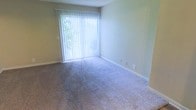 This single-story space features a well-proportioned room finished with wall-to-wall carpeting in a neutral gray-blue tone, creating a soft and understated foundation throughout. The walls are painted in a light, muted green-beige hue, contributing to a calm and functional atmosphere. A prominent floor-to-ceiling window fitted with vertical blinds serves as the room's focal point, drawing in generous amounts of natural light and offering a glimpse of lush outdoor greenery beyond. The blinds provide flexible light control while maintaining a clean, simple aesthetic. White baseboards frame the perimeter of the room, providing a crisp contrast against the neutral walls and carpet. Electrical outlets are conveniently positioned along the walls. The textured ceiling adds a subtle architectural detail consistent with the overall practical design of the space. The layout presents an open, versatile area that can accommodate a variety of furniture arrangements, making it adaptable for multiple uses. The combination of natural light, soft carpeting, and neutral tones gives the space a quiet, livable quality suitable for everyday use.
