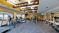 This fitness center in Tomball, TX offers a thoughtfully designed workout environment that blends rustic aesthetics with modern functionality. The space is defined by its striking ceiling, which features exposed dark wooden beam trusses set against yellow accent panels and lined with elongated fluorescent light fixtures, creating a bright and visually engaging atmosphere. Ceiling fans provide additional airflow throughout the facility.

The main workout area is generously sized and floored with durable, commercial-grade carpet tiles in a blue-grey tone. Large horizontal windows along one wall allow natural light to fill the space while offering views of the surrounding community. Mounted flat-screen TVs are positioned for convenient viewing during workouts.

The cardio zone is well-stocked with multiple treadmills, elliptical trainers, and stationary bikes arranged in an open row, ideal for sustained aerobic exercise. The strength training area includes a multi-station cable machine, a functional trainer, adjustable benches, and a full dumbbell rack. A dedicated stretching and functional fitness corner features medicine balls and balance training equipment. Wall-mounted mirrors in the strength area enhance the sense of space and allow for form monitoring. Fitness center rules are clearly posted near the entrance, ensuring a structured and safe environment for all users.