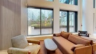 Located at 422 Cherry Street in Toronto's vibrant waterfront community, this contemporary lobby lounge is a standout communal living space defined by its striking architectural design. The room commands attention with dramatic double-height ceilings and expansive floor-to-ceiling windows framed in sleek black steel, flooding the interior with natural light and offering unobstructed views of a beautifully landscaped courtyard. Large-format light stone tile flooring extends across the entire level, lending a clean, polished aesthetic throughout. A sculptural staircase with a minimalist black metal railing anchors one end of the space, contributing to the verticality of the room. Warm wood-paneled accent walls provide a refined contrast to the crisp white surfaces, while curated artwork — including a bold blue canvas and a vibrant multicolored piece — adds cultural character. The living area is thoughtfully arranged with plush earth-toned sofas, contemporary lounge chairs in cognac leather, and a collection of bistro-style tables and chairs ideal for casual gathering or focused work. A glass-partitioned meeting room with pendant lighting is accessible via full-height black-framed doors, offering a more private setting within the broader space. The overall ambiance is modern, welcoming, and professionally curated.