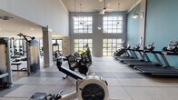 This fitness center is designed to accommodate a wide range of workout styles within a generously proportioned, single-level space. The main cardio area is immediately striking, featuring soaring double-height ceilings lined with large, grid-paned windows that flood the room with natural light and offer views of the surrounding exterior. Ceiling fans and pendant lighting complement the bright, airy atmosphere. Treadmills, ellipticals, stationary bikes, and rowing machines are thoughtfully arranged throughout this zone, providing ample room between equipment.

Moving deeper into the facility, structural columns define distinct workout zones without obstructing the open feel of the space. A dedicated strength area houses a full dumbbell rack, cable machines, and selectorized weight stations, with mirrored walls enhancing the sense of depth. A wall-mounted television provides entertainment throughout this section.

A separate functional training room features teal accent walls, wall sconces, and a mini-split climate system. This zone is equipped with kettlebells, battle ropes, TRX suspension trainers, medicine balls, and a BOSU balance trainer, offering a complete space for dynamic and circuit-style workouts. Large-format tile flooring with rubber sport inlays runs throughout, providing durability and safety across all zones.