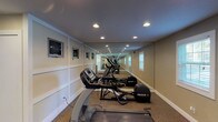 This single-floor community facility offers a thoughtfully designed collection of spaces that blend comfort and functionality. The welcoming reception area greets visitors with soaring ceilings, recessed lighting, and a striking multi-bulb chandelier. Dark-framed double entry doors allow abundant natural light to flood the space, complemented by neutral tones and tasteful décor including floral arrangements and curated artwork.

The lounge area is a standout, featuring a dramatic floor-to-ceiling stone fireplace with a rustic wood mantel, creating a warm and inviting focal point. Comfortable seating, including plush sofas and wingback chairs in soft neutral hues, is arranged throughout, while black-framed French doors open to an outdoor area, enhancing the sense of openness. A mounted flat-screen television adds a modern amenity to the relaxed setting.

The fitness center is well-equipped with a lineup of cardio machines, including treadmills and ellipticals from leading brands. Full-length mirrors line one wall, visually expanding the space and supporting workout visibility. Wall-mounted televisions and recessed lighting complete the practical, clean environment. Together, these areas create a versatile and welcoming community hub designed for both relaxation and active use.